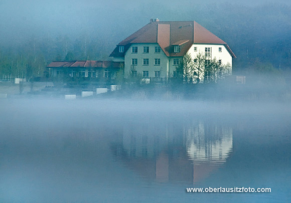 Foto von Peter Hennig PIXELWERKSTATT Das Hotel Haus am See an einem nebligen Morgen. Das Haus spiegelt sich im Wasser.